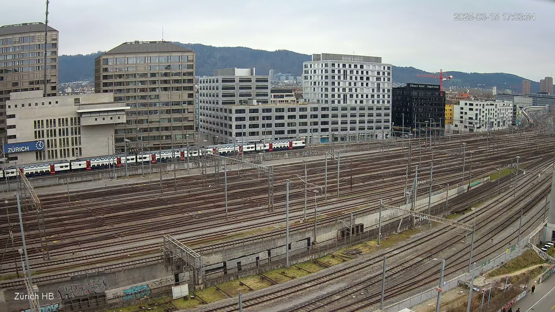 A long expanse of train tracks stretches out before several multi-story buildings under an overcast sky.