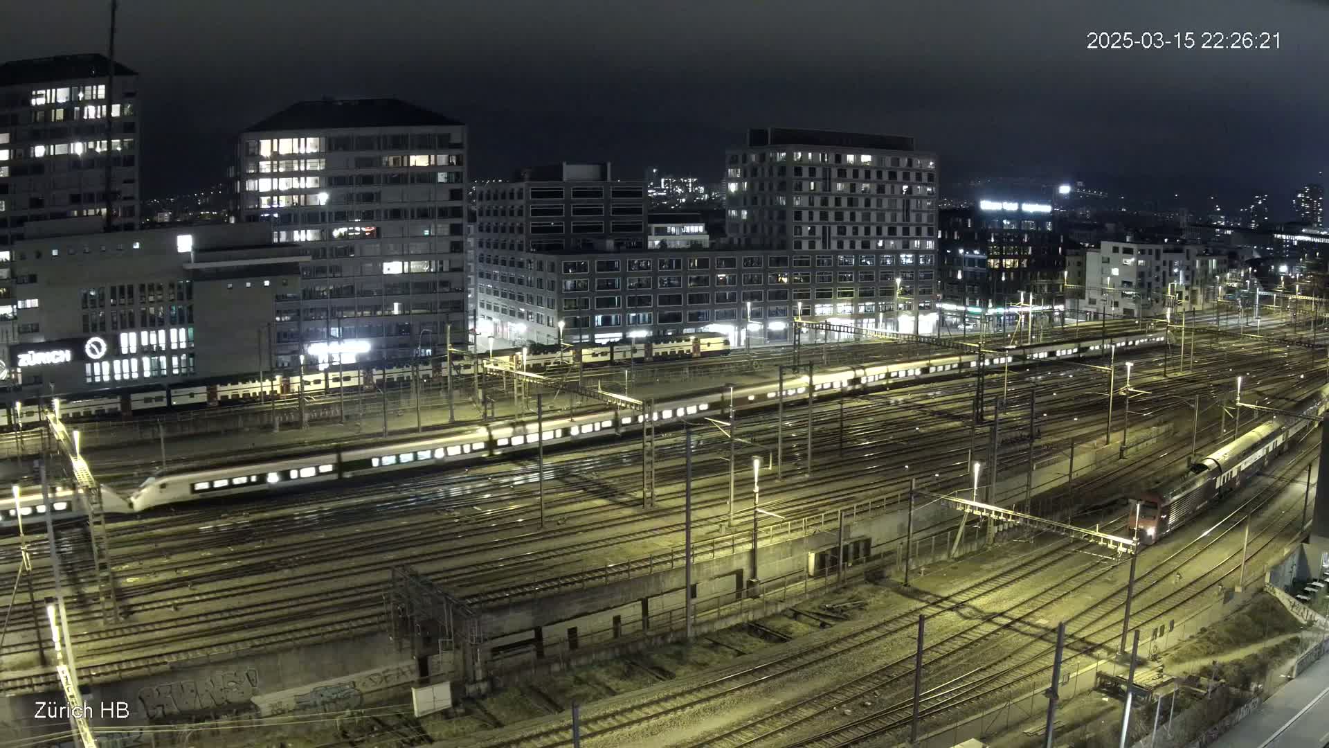 A nighttime aerial view of a sprawling railway yard with several trains, surrounded by brightly lit city buildings under a dark sky.
