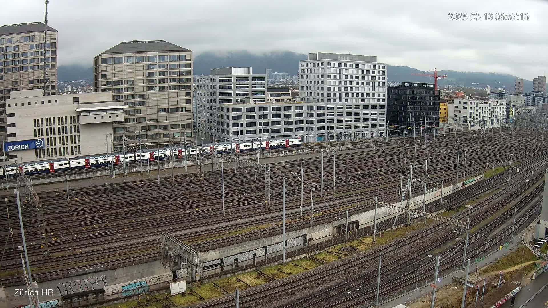 A long expanse of train tracks sits below a cluster of modern and older buildings on a cloudy day.