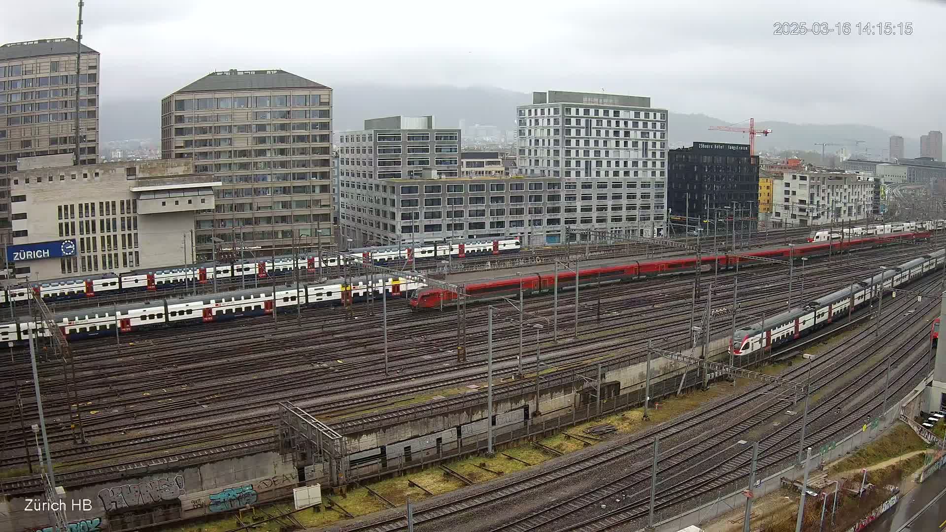 A wide shot of a train yard with many trains and city buildings under an overcast sky.