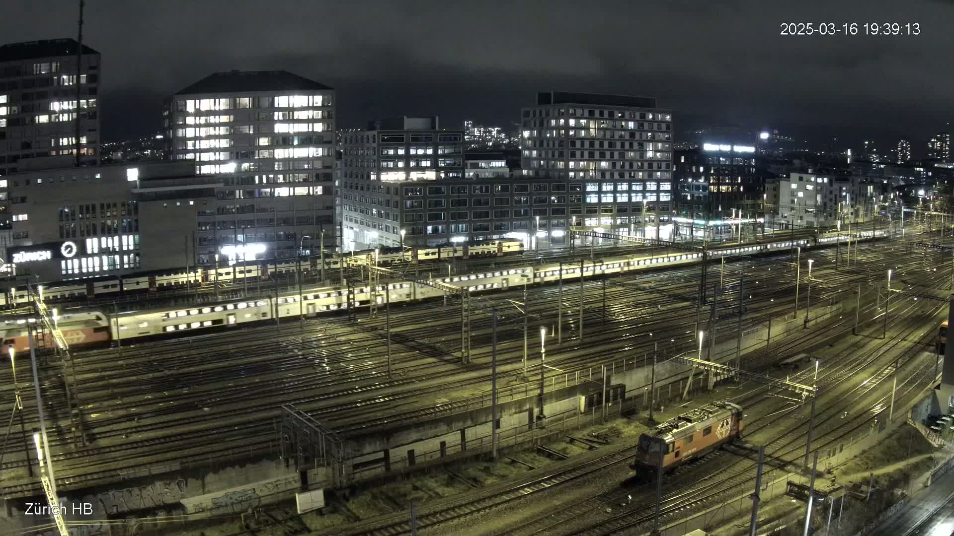 A nighttime view of a large train yard with multiple passenger trains and a locomotive, surrounded by illuminated buildings under a dark, cloudy sky.
