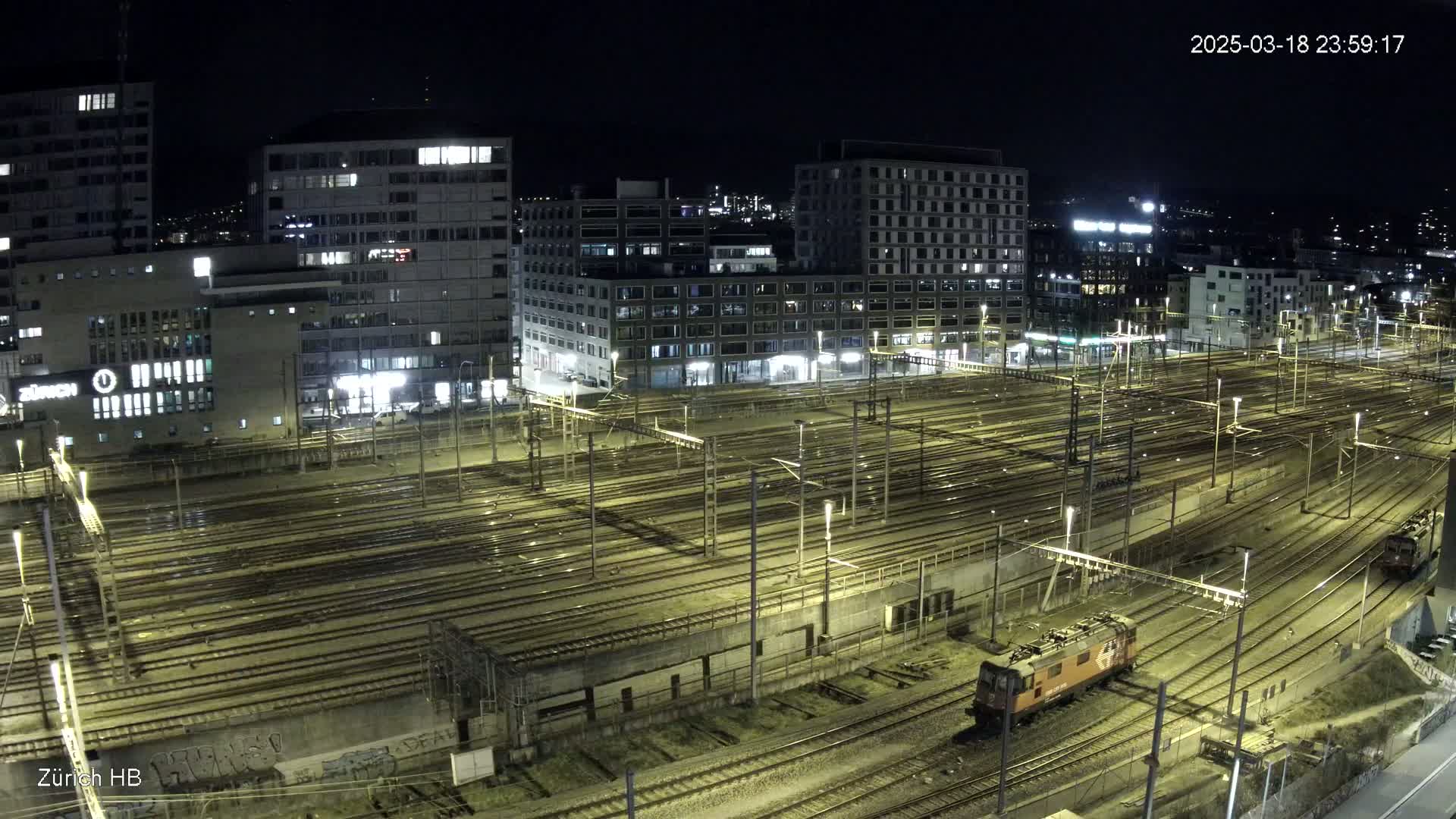 A nighttime, high-angle view of a large train yard with many tracks and buildings illuminated, showing a single locomotive on one of the tracks.