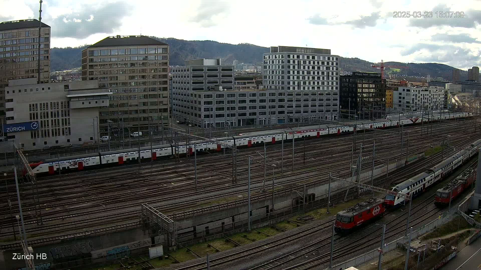 A long passenger train sits in a train yard amongst numerous train tracks and modern buildings under a partly cloudy sky.