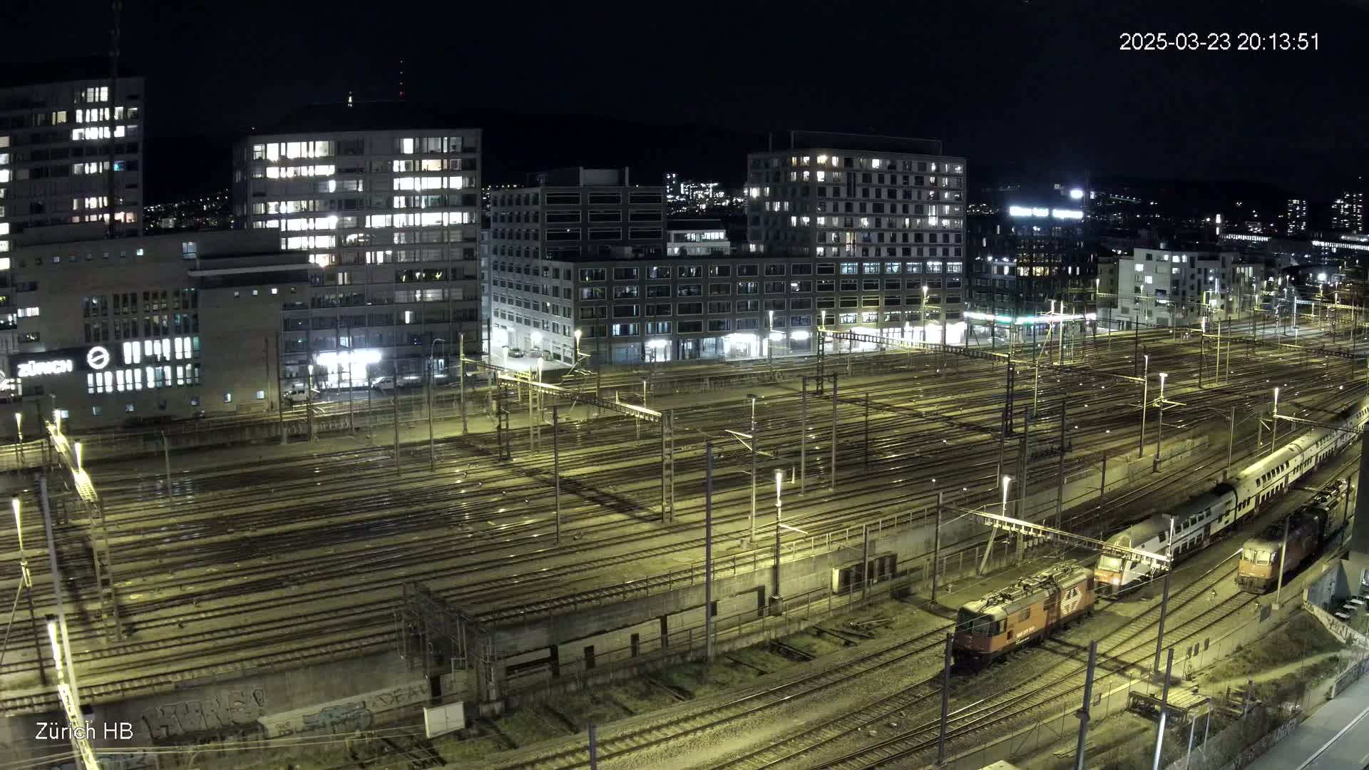 A nighttime, high-angle view of a large, illuminated train yard surrounded by multi-story buildings, with several trains visible on the tracks.