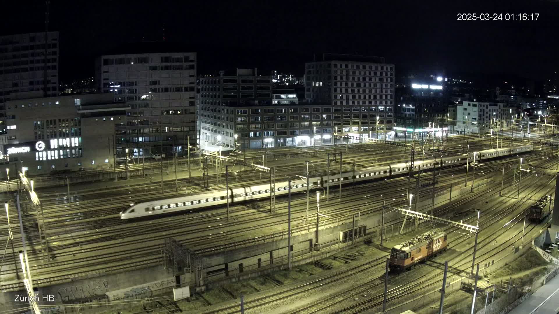 A long, white passenger train moves across a complex network of illuminated train tracks at night, with several buildings visible in the background.