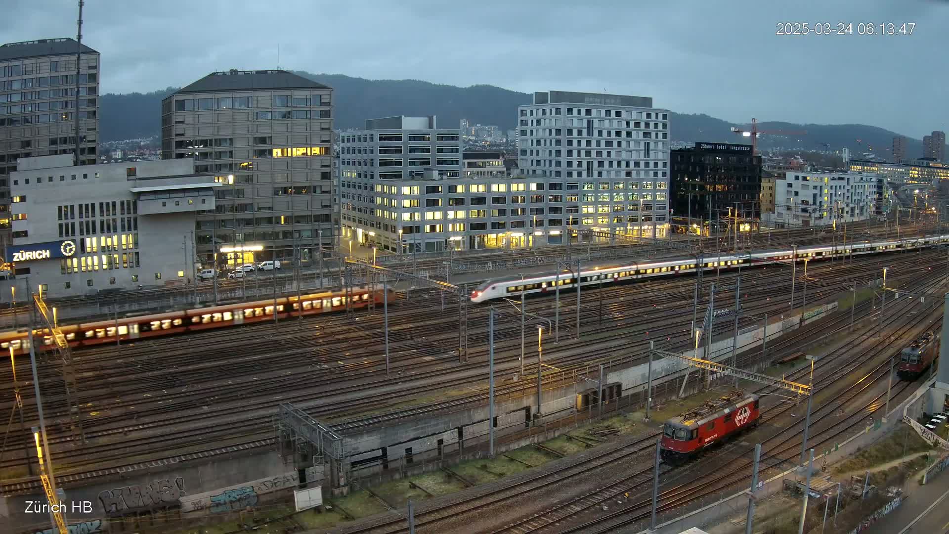 A high-angle, long shot of several trains on a complex railway system next to a city under an overcast sky.