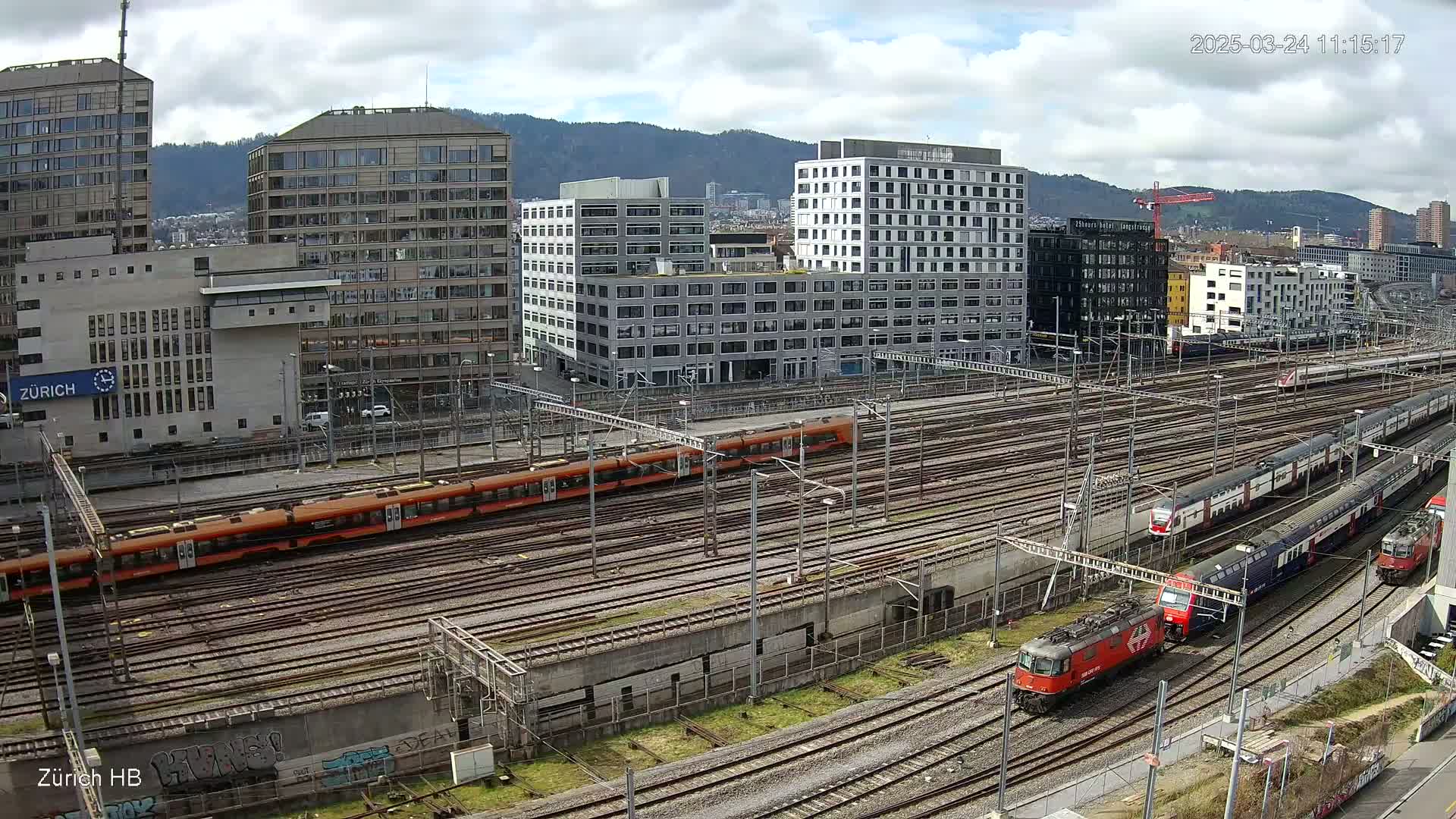 A high-angle view of a train station with multiple trains on numerous tracks, under a partly cloudy sky, surrounded by modern buildings and mountains in the distance.