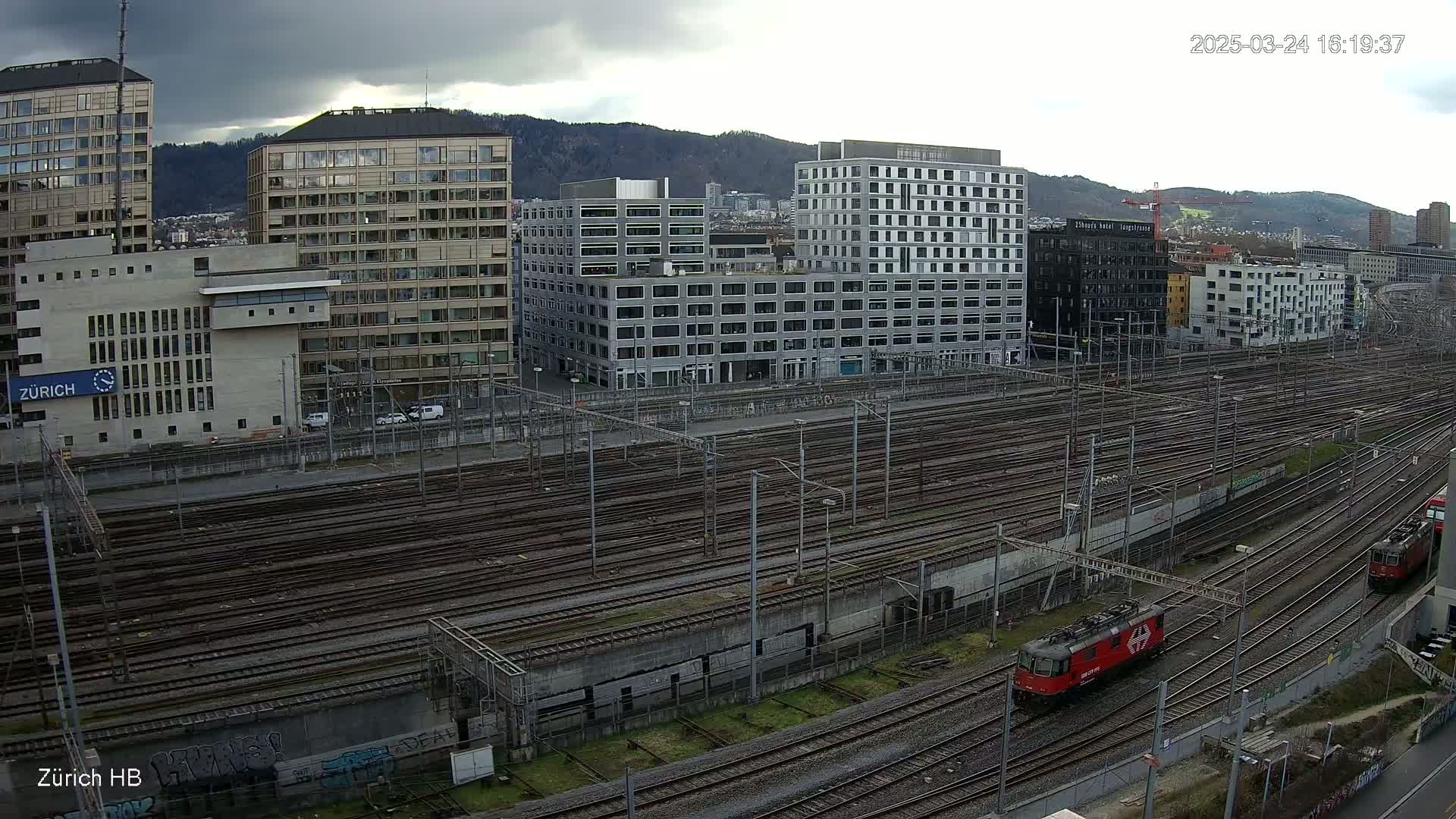 Under a cloudy sky, a red train travels along numerous railway tracks beside several multi-story buildings.
