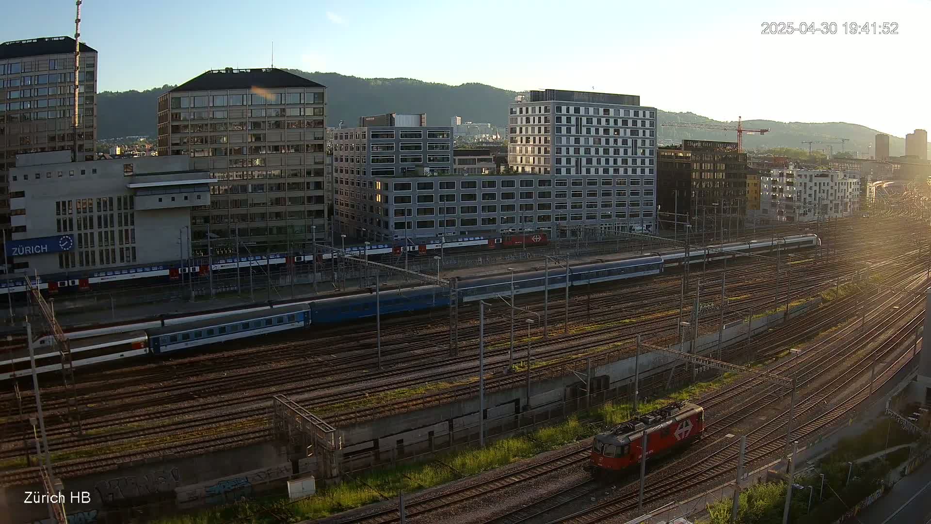 A long passenger train and a red locomotive are on a sprawling railway network next to several modern and older buildings under a clear, sunny sky.