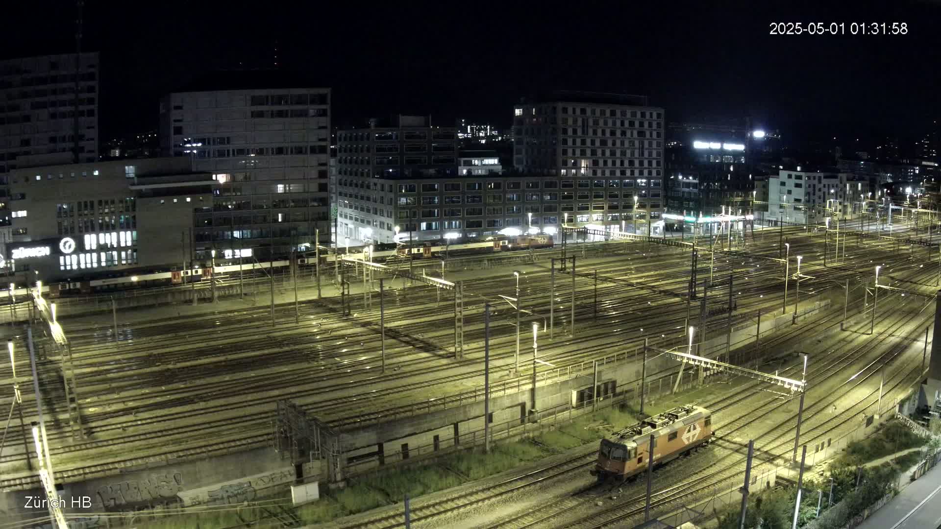 A nighttime, high-angle view of a sprawling railway yard with numerous train tracks, surrounded by city buildings.