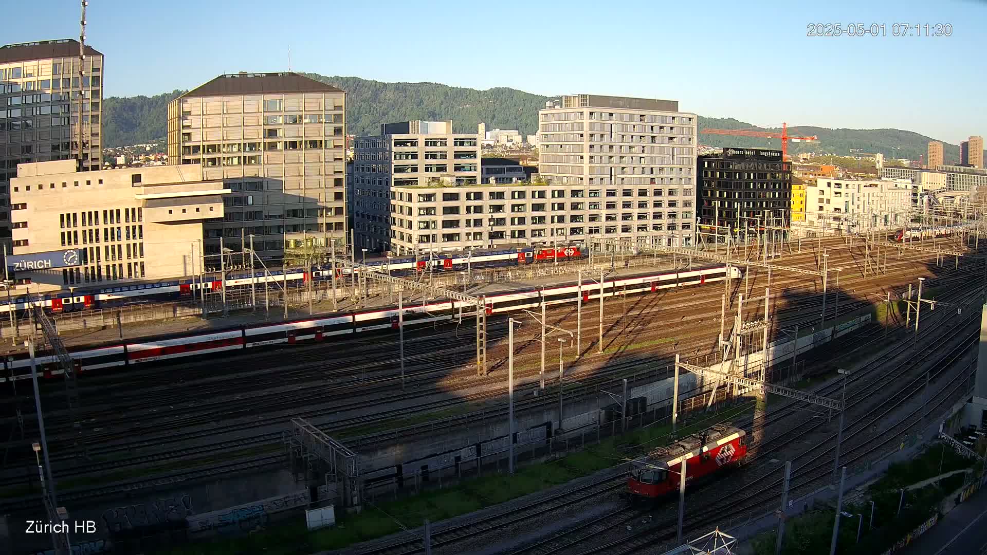 Several trains are parked on a sprawling network of train tracks near a cluster of modern buildings under a clear, sunny sky.