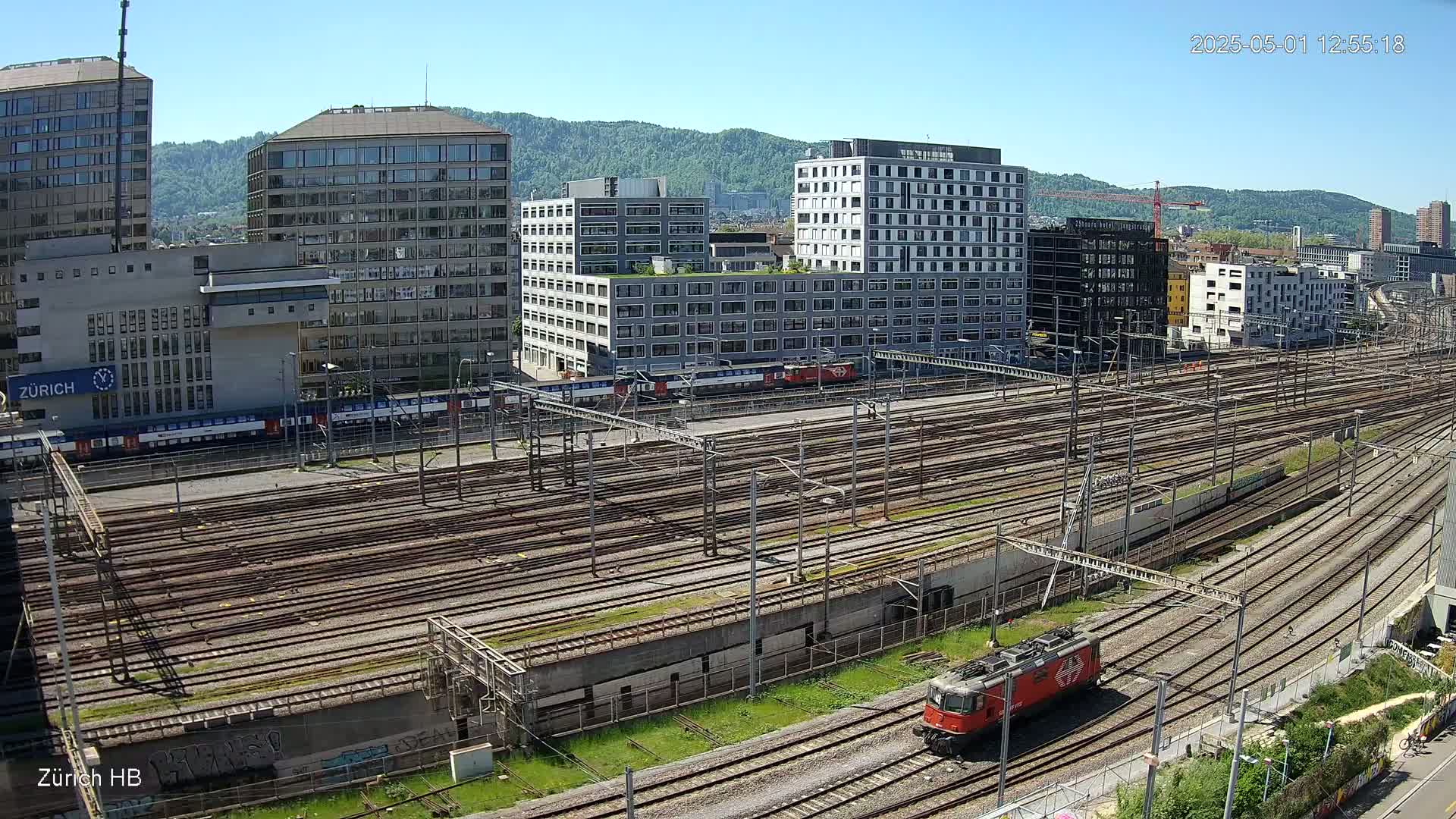 A wide, high-angle view shows a sprawling railway yard with numerous train tracks under a clear, sunny sky, surrounded by modern buildings and lush green vegetation.