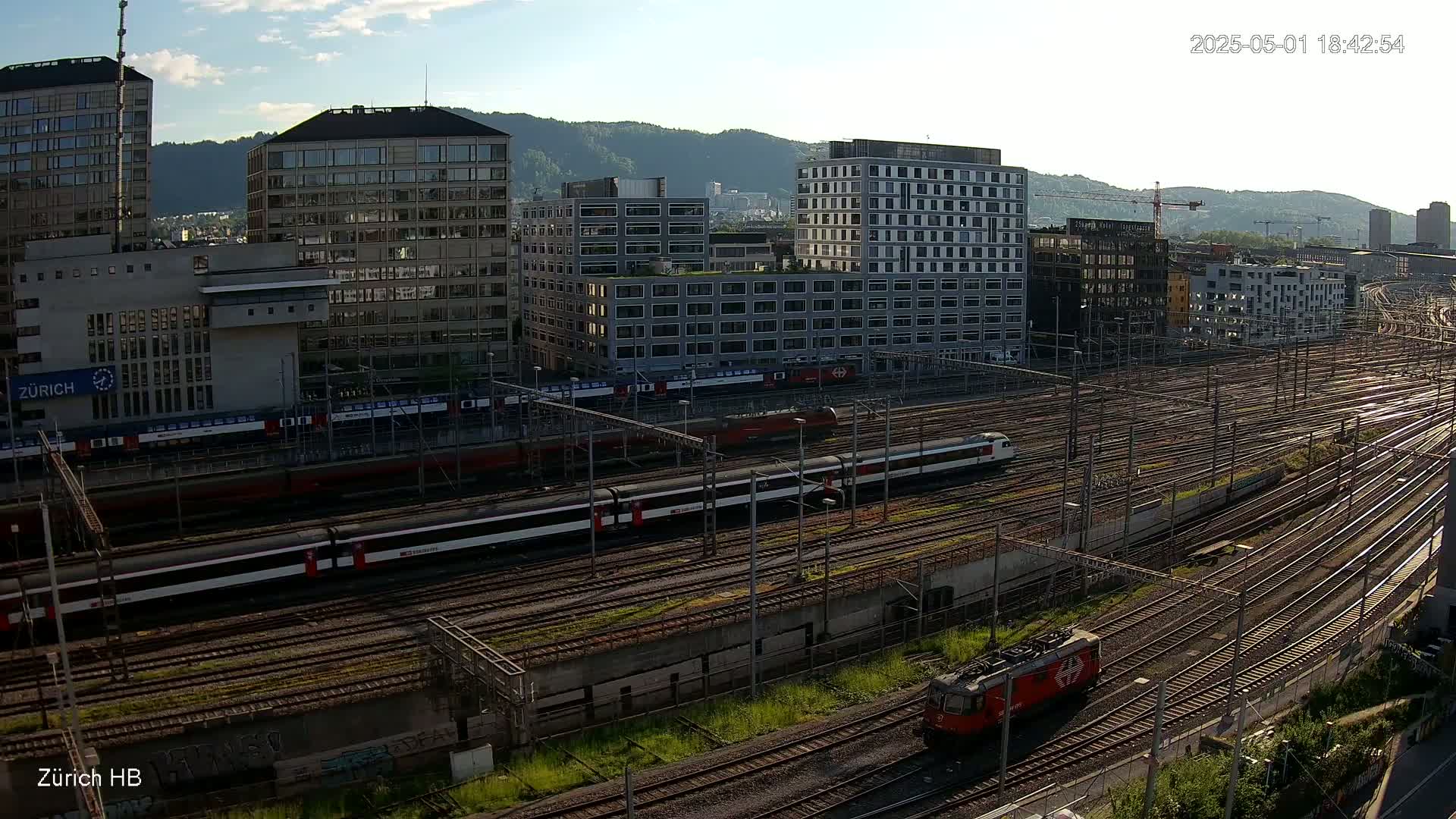 A sunny day view of a large train station with multiple trains on numerous tracks, surrounded by modern buildings and hills in the background.