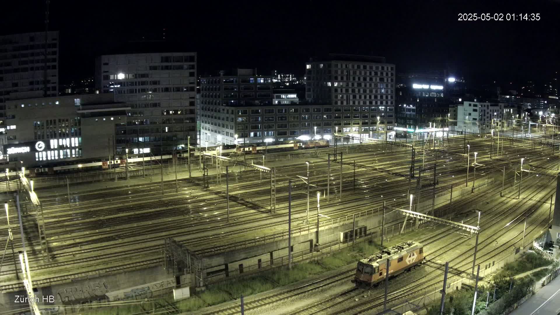 A nighttime aerial view shows a large, illuminated train yard with numerous tracks and several buildings in the background under a clear sky.