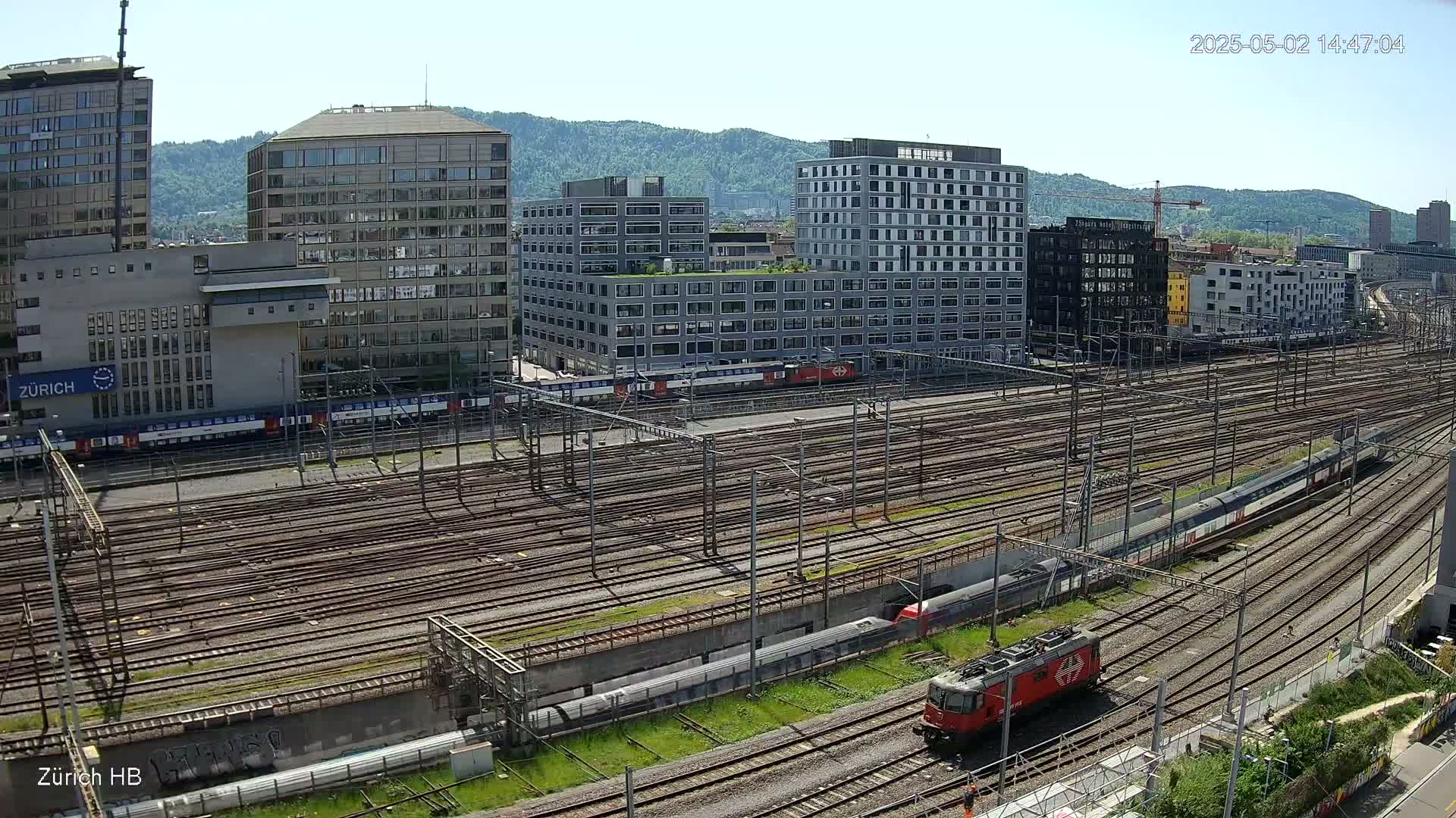 A high-angle, sunny view of a large railway yard with multiple train tracks, surrounded by modern buildings and hills in the background.