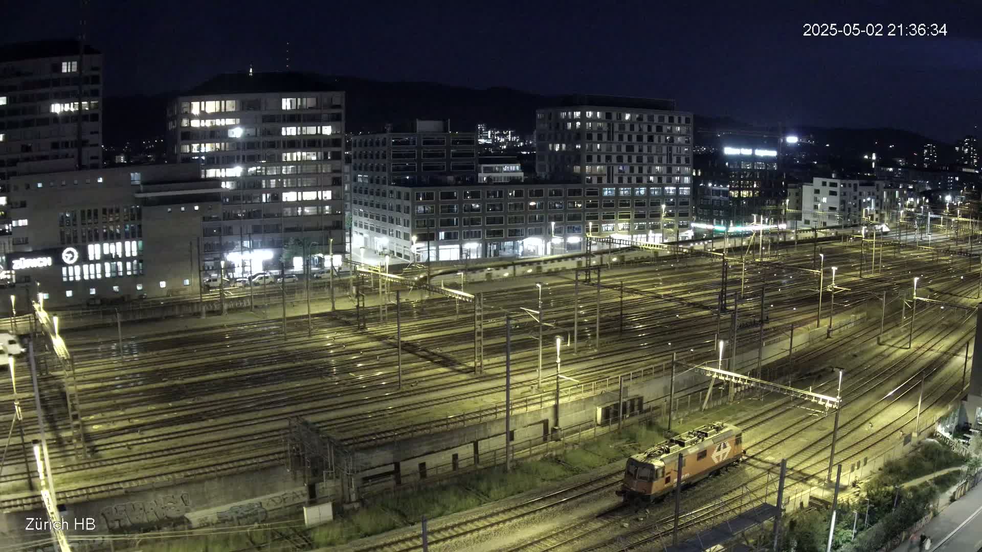 A nighttime, high-angle view of a train yard with numerous tracks and lit buildings in the background.