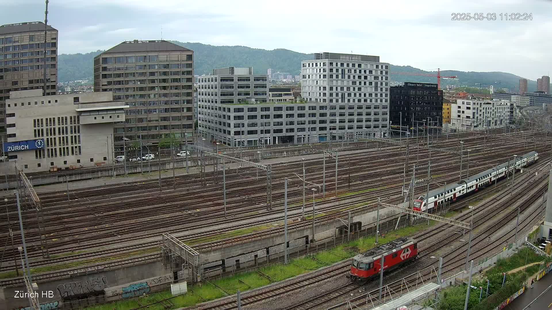 A view of a railway yard with numerous tracks and modern buildings under an overcast sky, with two trains visible.