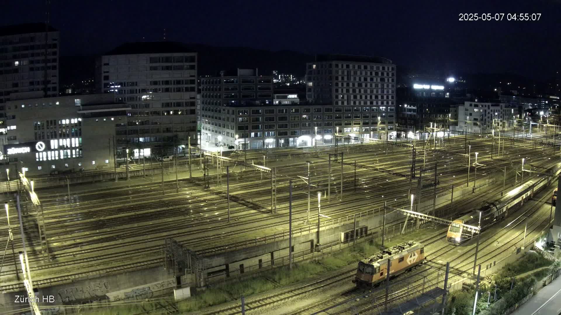 A nighttime, high-angle view shows a large train yard with many tracks, illuminated by lights, and two trains; one is an orange locomotive and the other is a long passenger train.