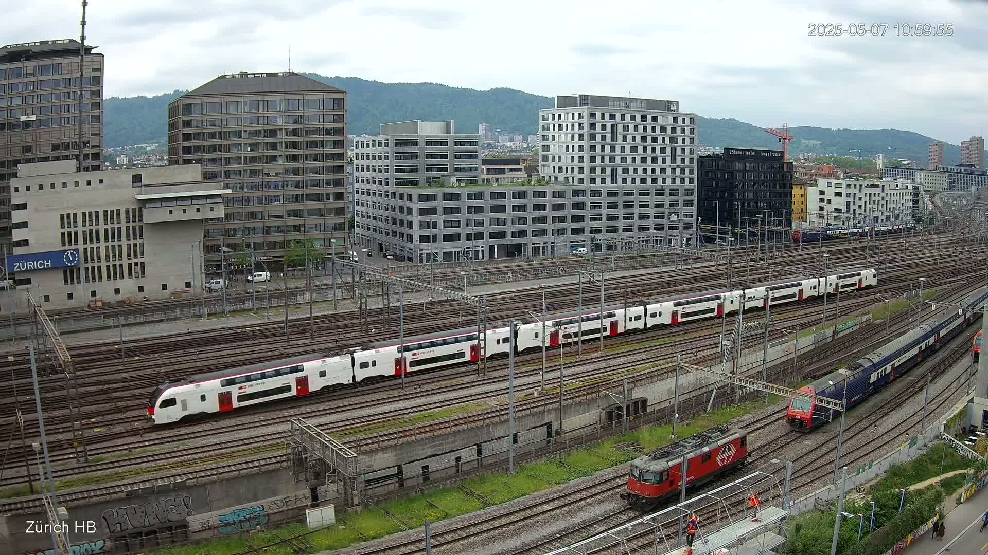 A long passenger train and a shorter freight train are visible on a railway network surrounded by modern buildings under an overcast sky.