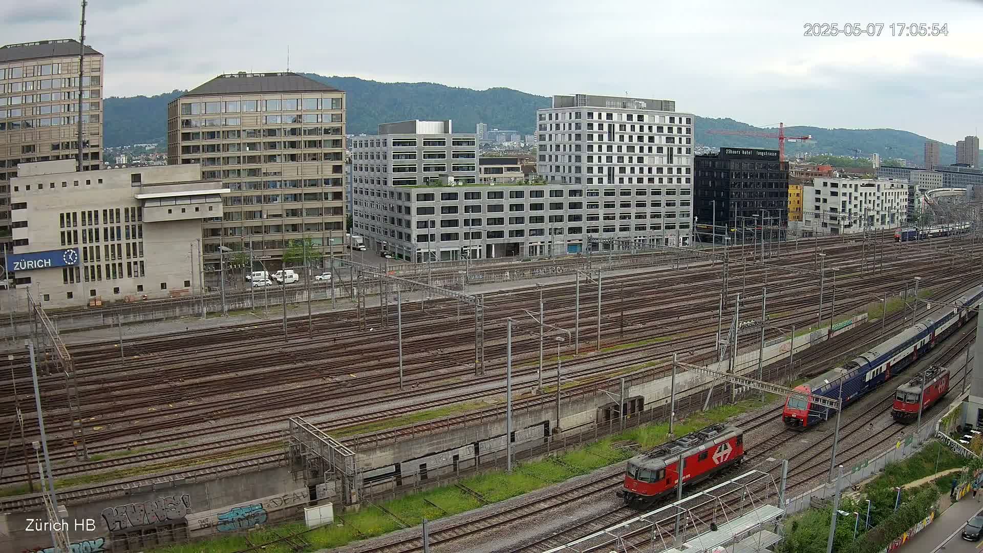 Two trains are traveling on a large network of train tracks beside several multi-story buildings under an overcast sky.