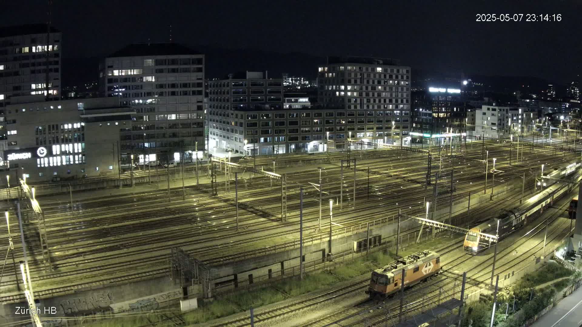 At night, two trains are seen on a large network of illuminated train tracks next to several city buildings.