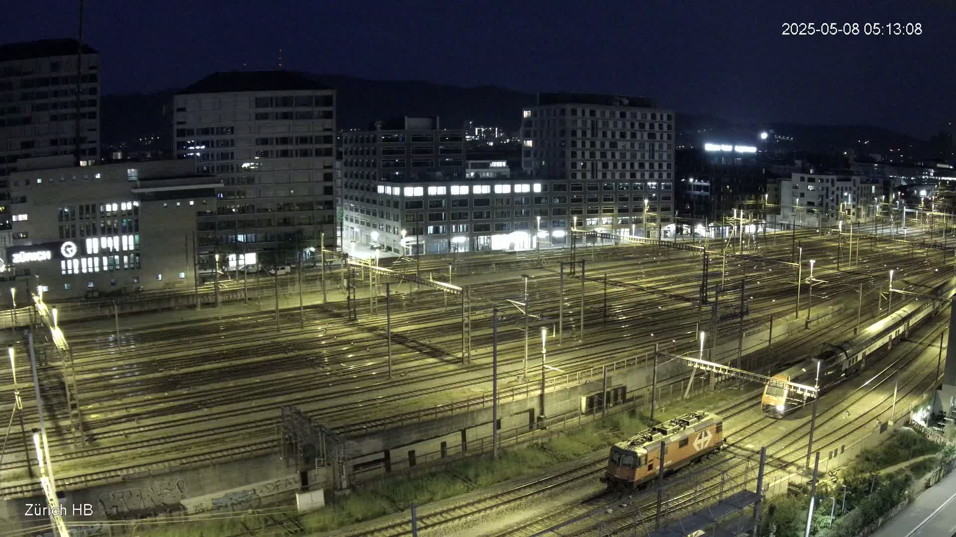 A nighttime view of a large train yard with multiple tracks and several trains, surrounded by illuminated buildings under a dark sky.