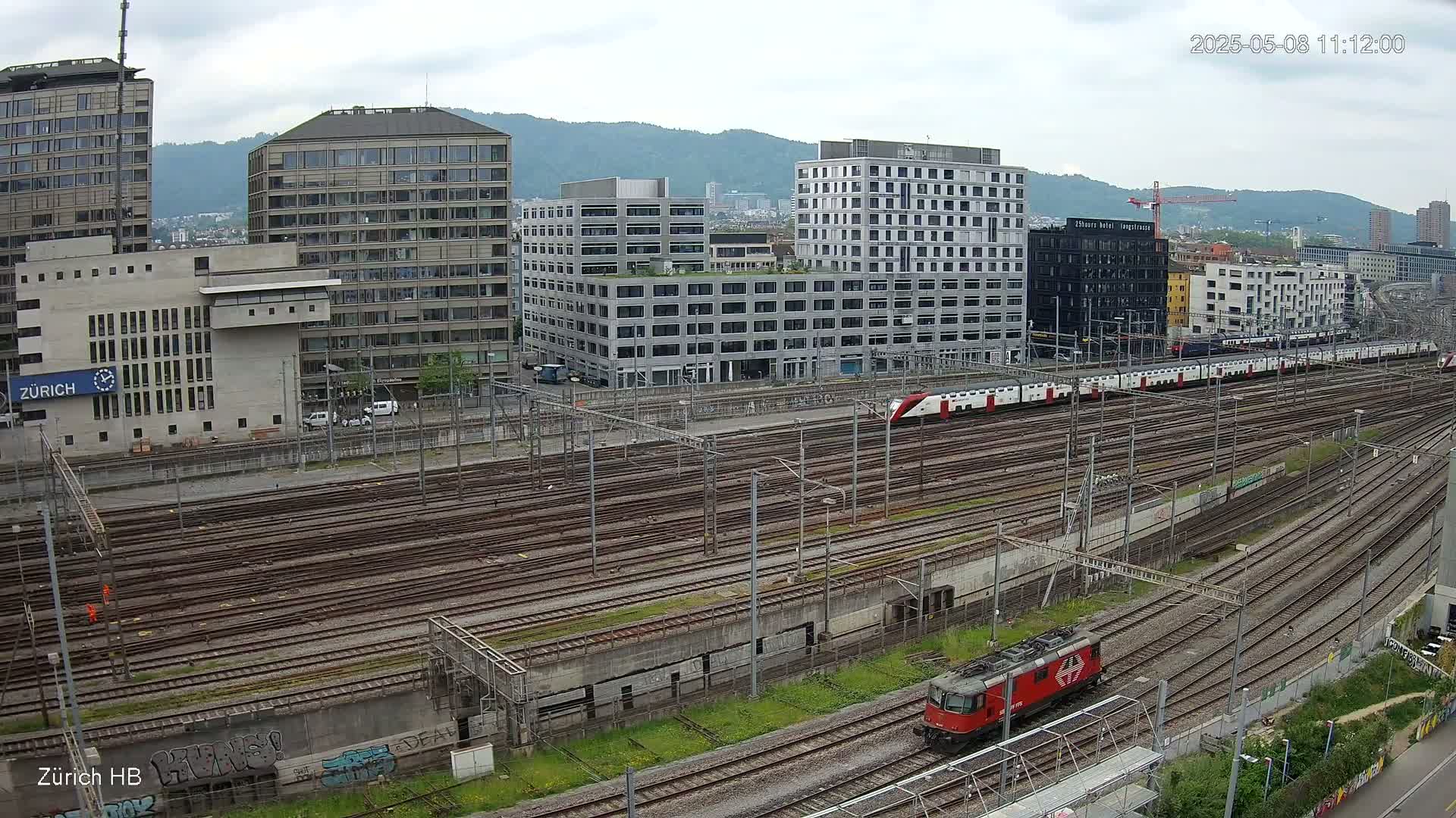 A high-angle view of a train station on an overcast day shows numerous train tracks with two trains, one long passenger train and one locomotive, and several modern buildings in the background.