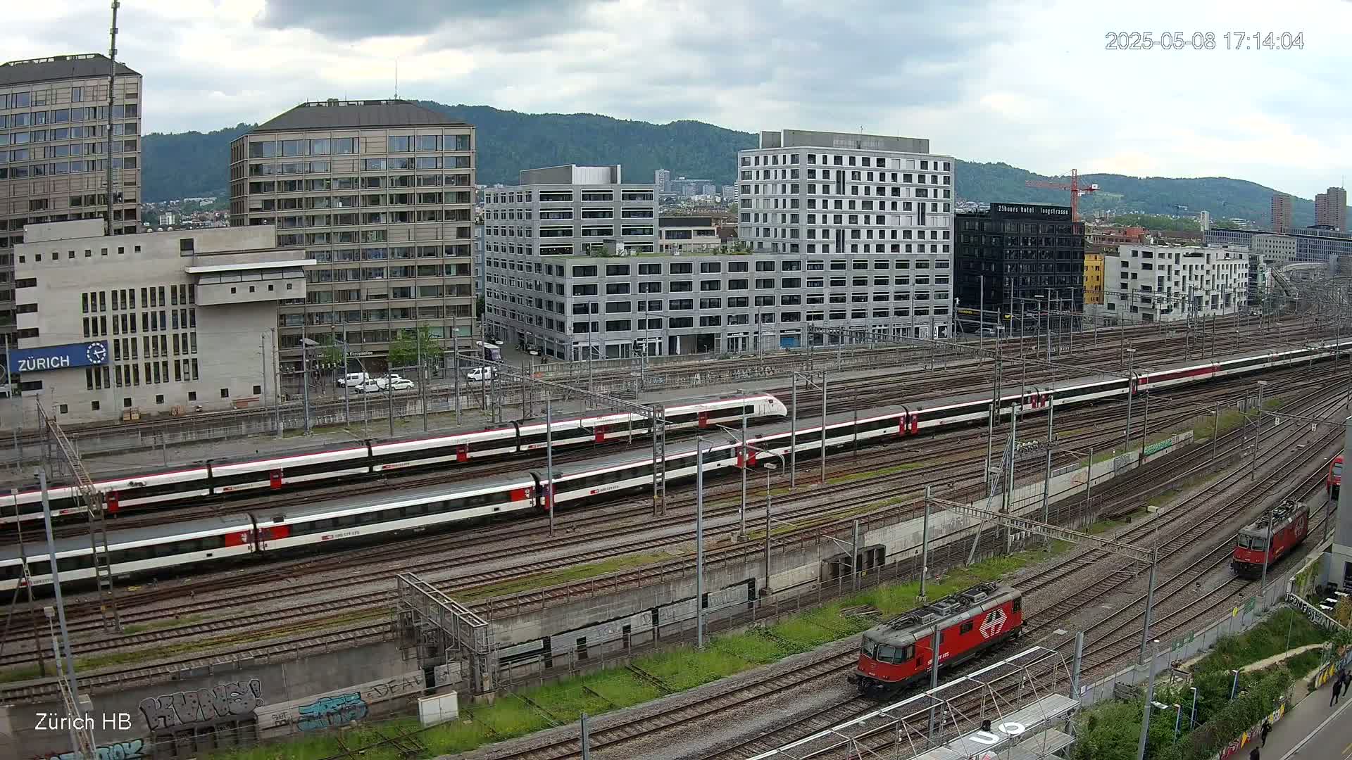 A high-angle view shows several trains on a railway network, surrounded by city buildings under a cloudy sky.