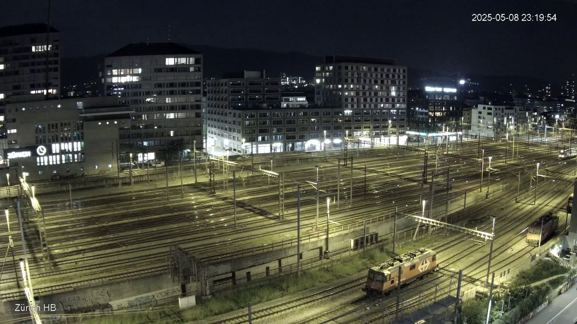 A nighttime view of a large, illuminated train yard with multiple tracks and buildings in the background.