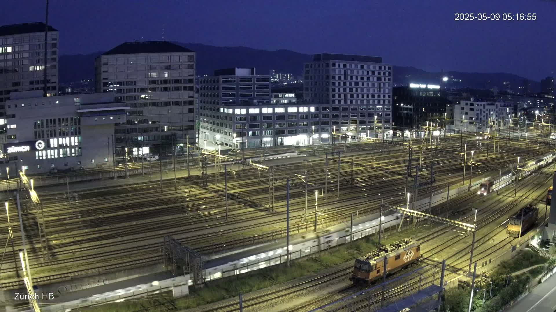 A nighttime view of a large train yard with multiple tracks, surrounded by city buildings under a dark, clear sky.