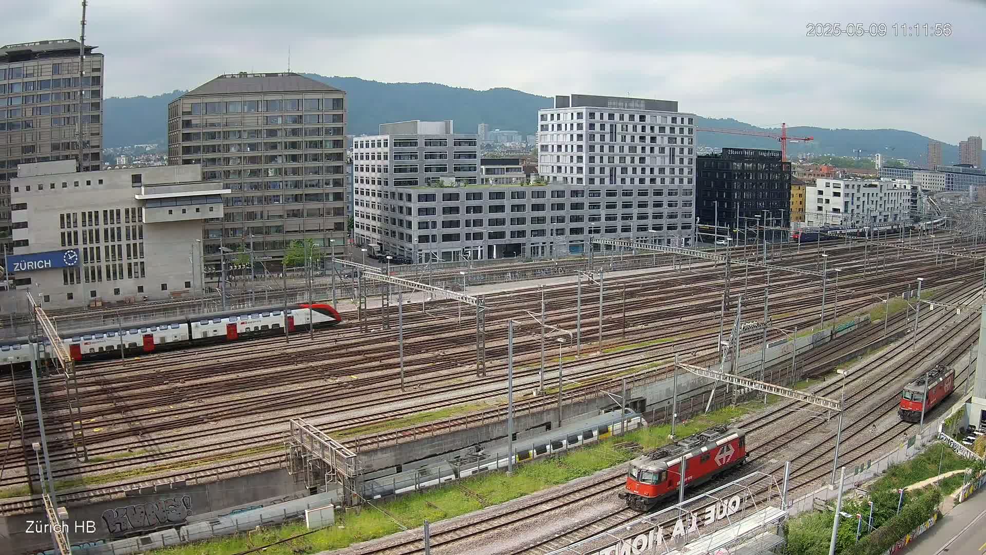 A view of a train station with many train tracks, multiple buildings, and two red locomotives under an overcast sky.