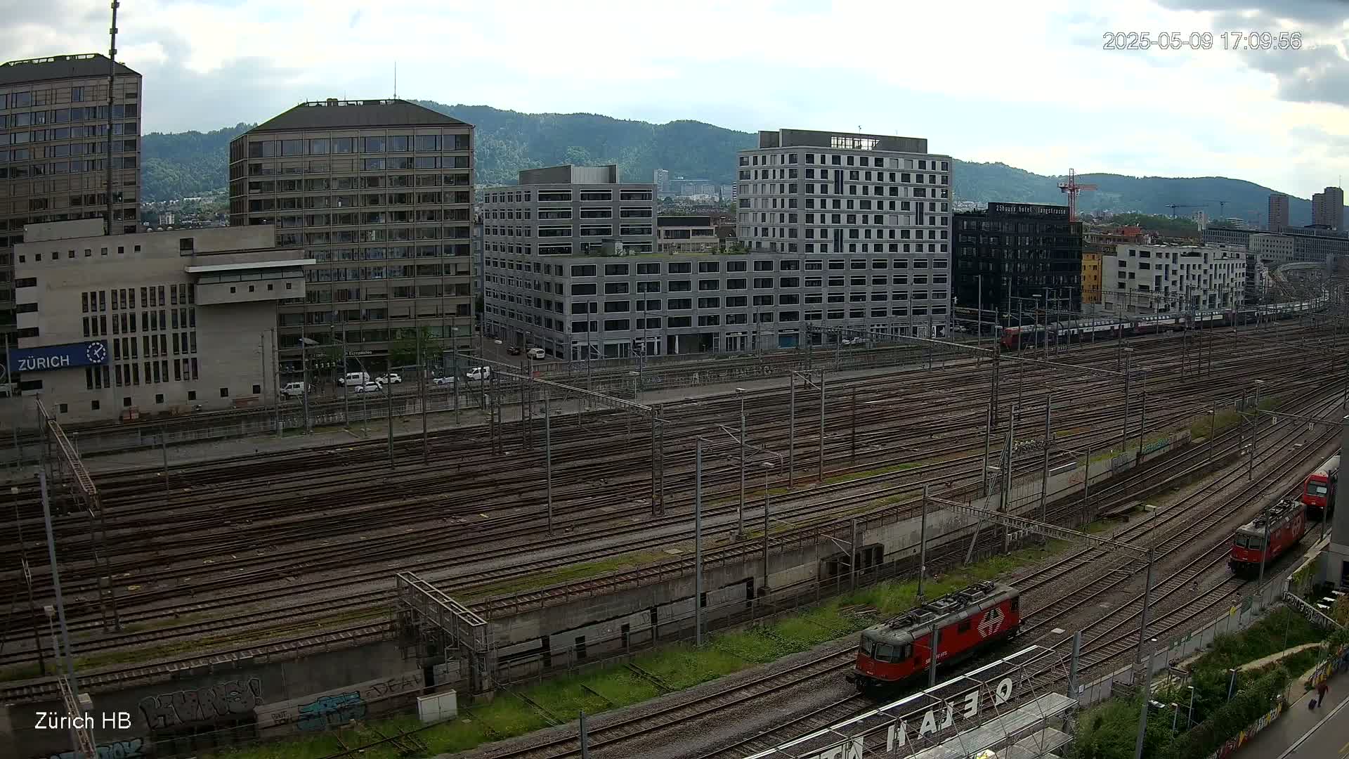 A high-angle view of a railway yard with numerous tracks, surrounded by modern buildings under a partly cloudy sky.