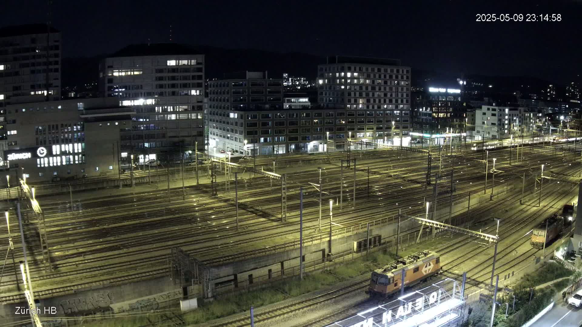 A nighttime aerial view of a large train yard with numerous tracks illuminated, surrounded by city buildings.