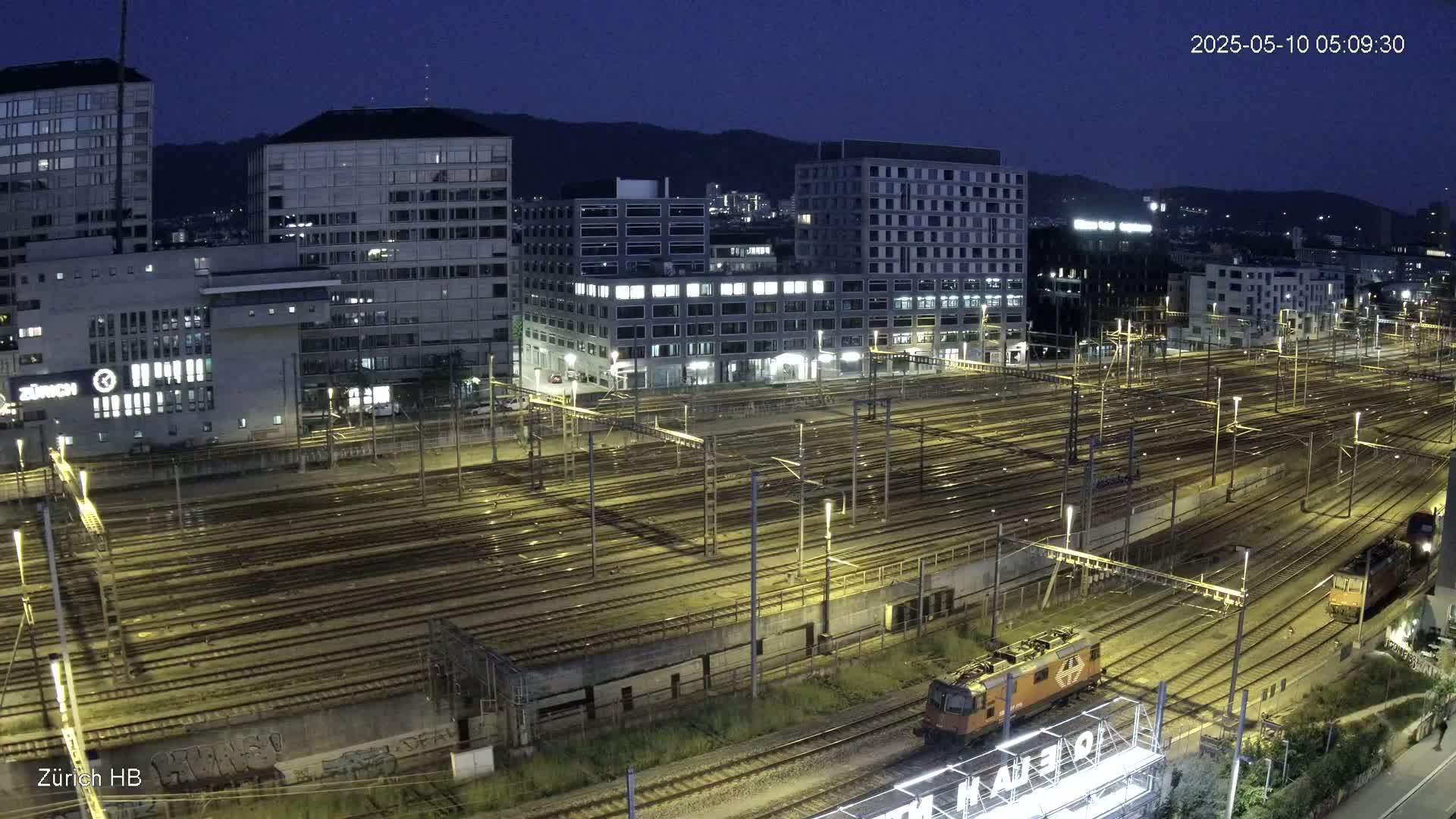 A nighttime, high-angle view of a large train yard with numerous tracks surrounded by city buildings under a dark sky.