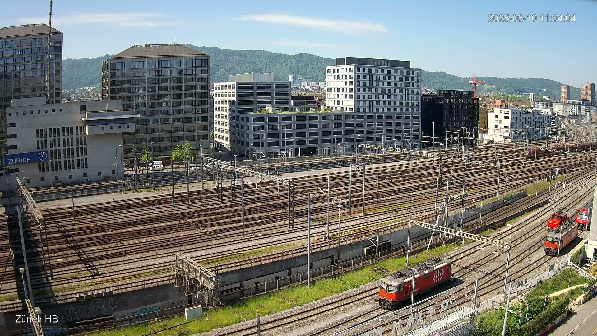 A sunny day reveals a large railway yard surrounded by modern buildings, with several red locomotives visible on the tracks.