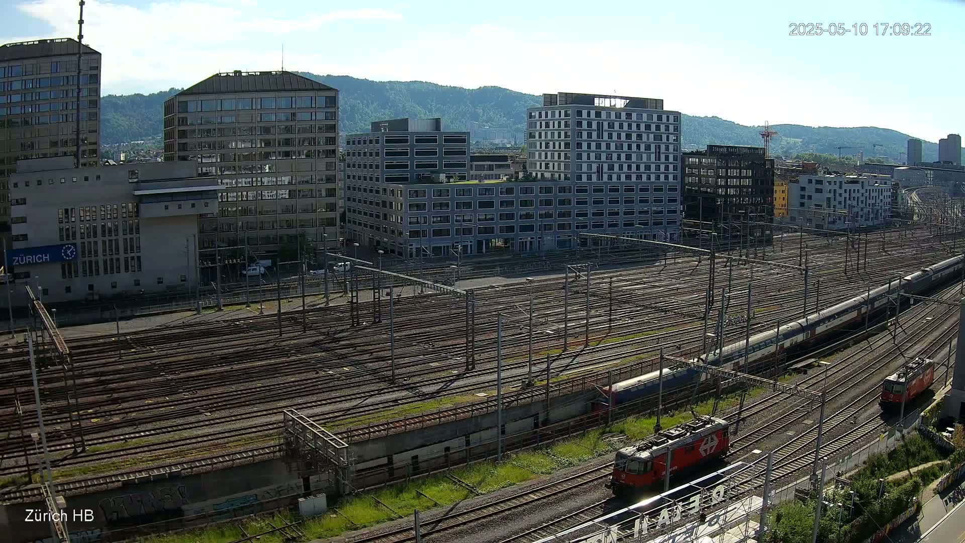 A high-angle view on a sunny day shows a large train yard with multiple tracks and several trains, surrounded by modern buildings and hills in the background.