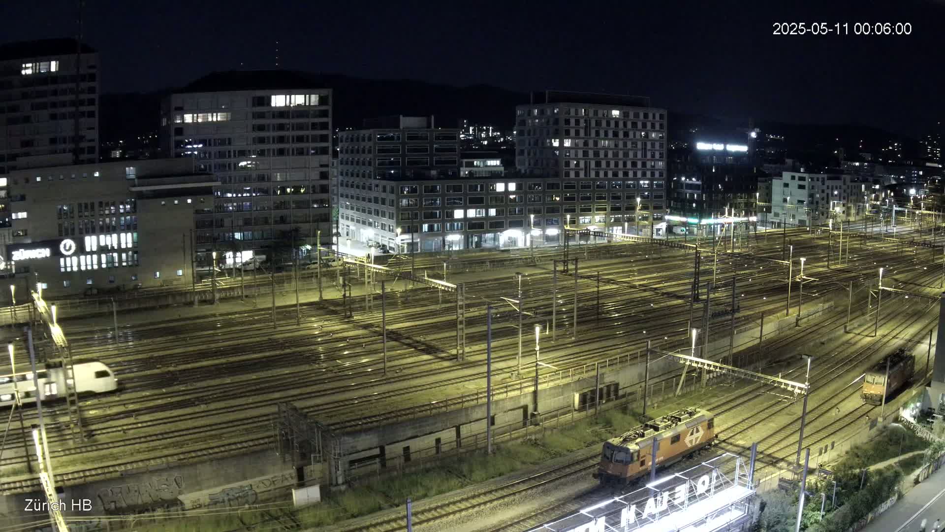 A nighttime, overhead view of a large train yard with multiple tracks, illuminated by lights, surrounded by buildings.
