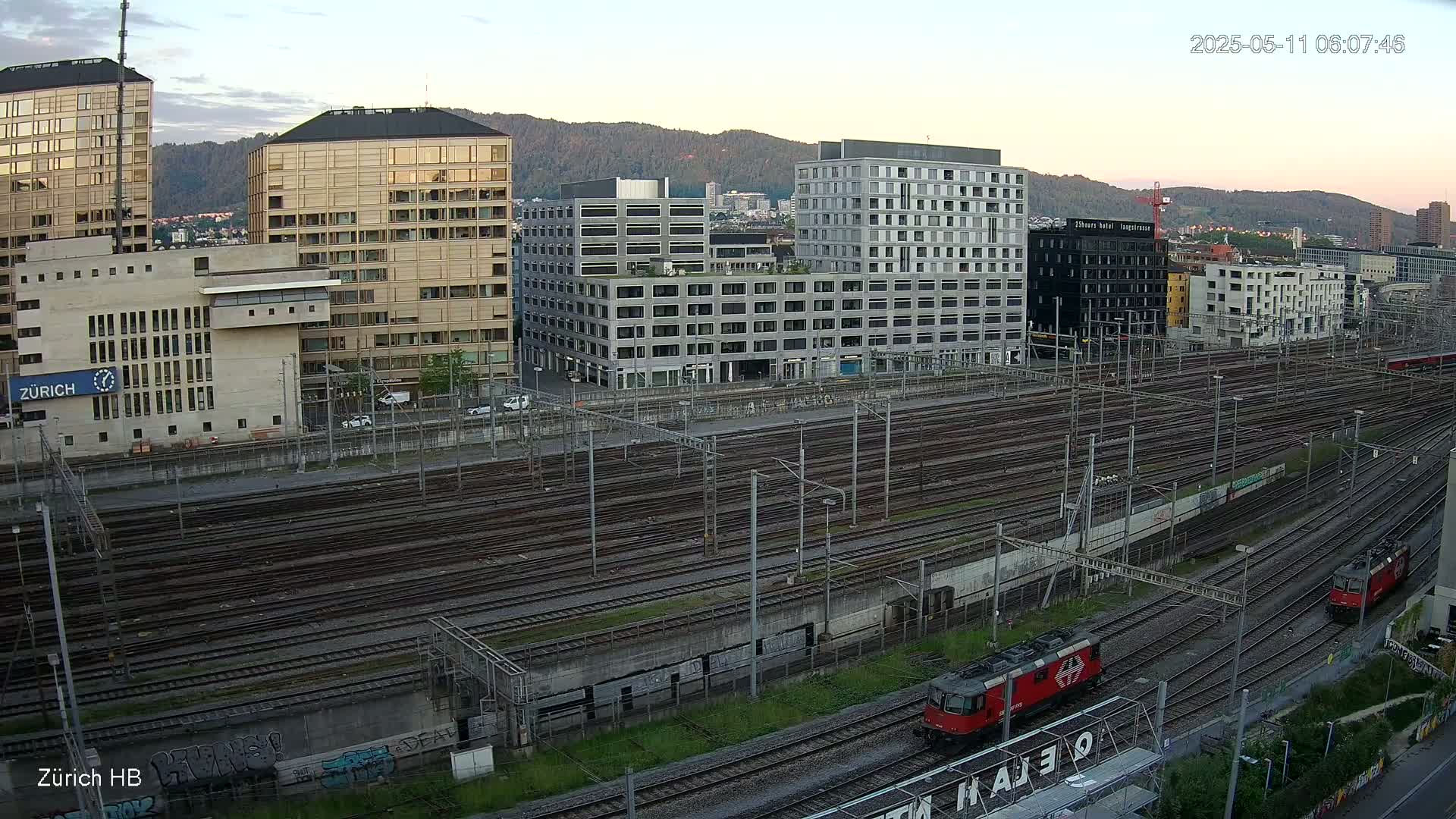 Two red locomotives are on a network of train tracks next to several modern buildings under a twilight sky.