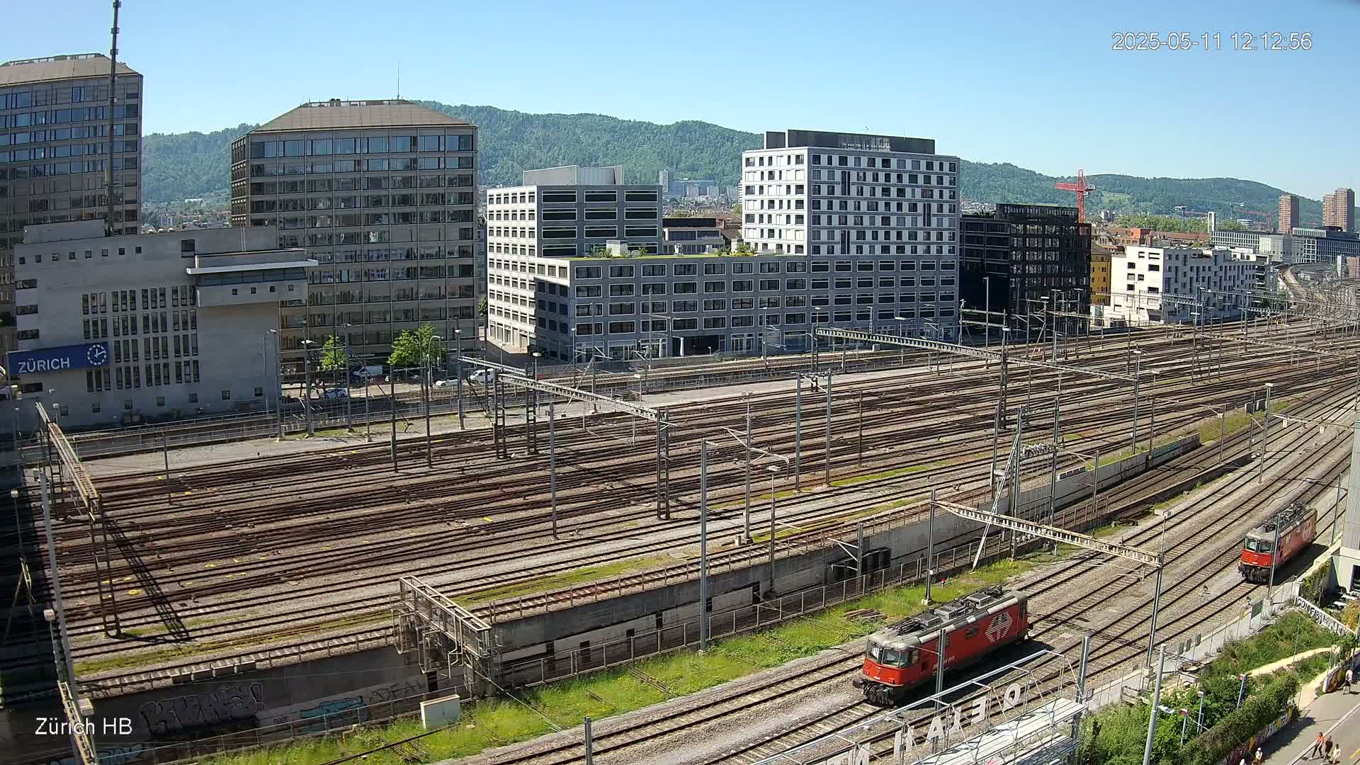 Two red locomotives are on a large, expansive train yard surrounded by modern buildings under a clear, sunny sky.