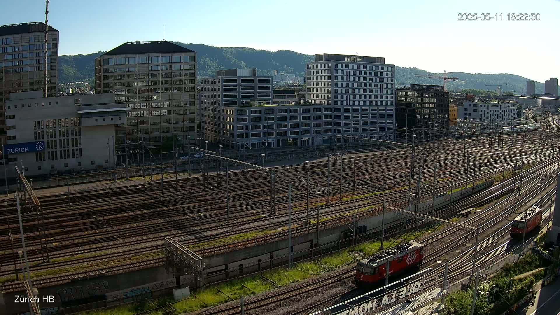 A large railway yard with numerous train tracks and several modern buildings in the background under a clear, sunny sky.