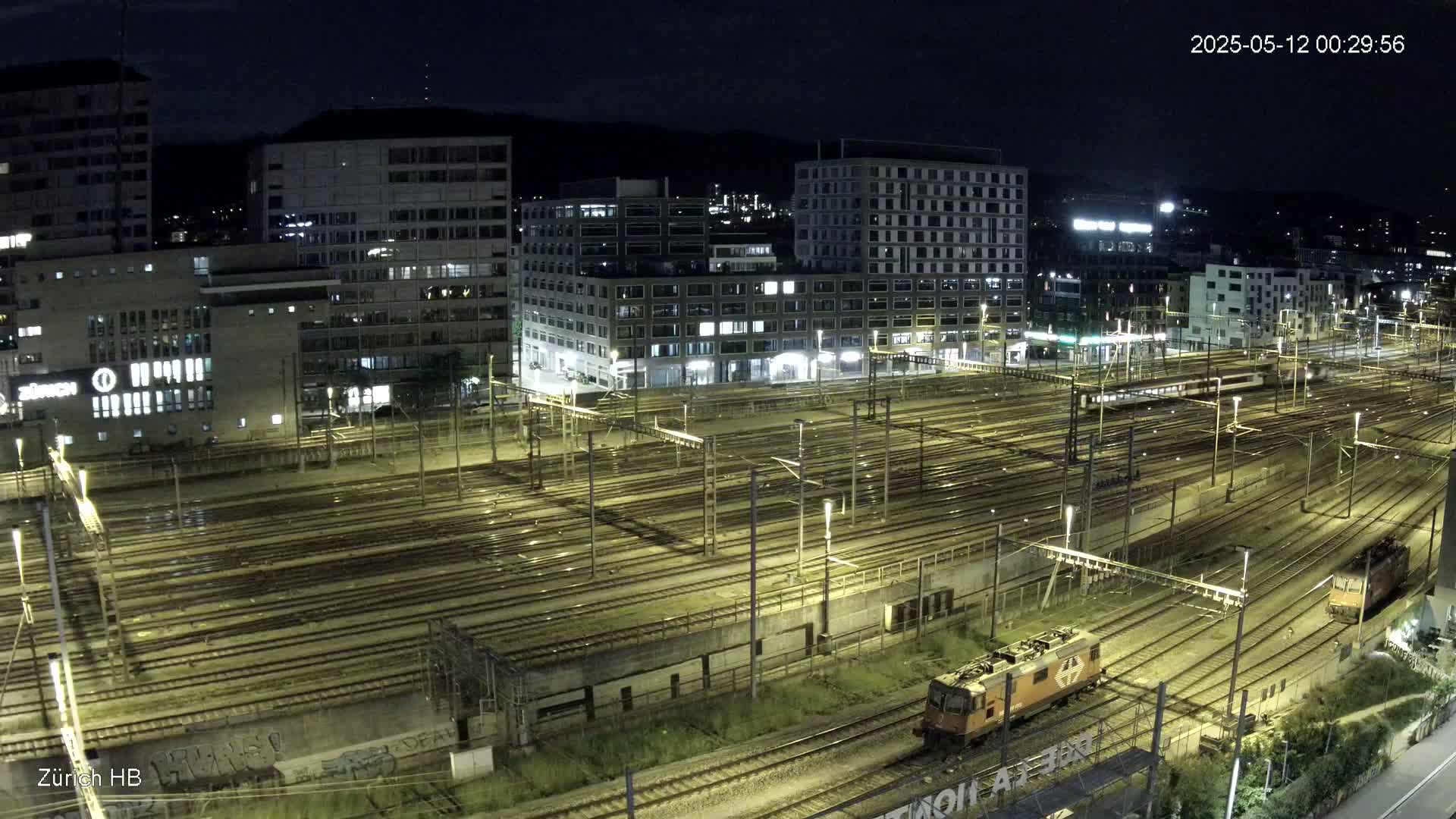 A nighttime, high-angle view of a sprawling train yard illuminated by lights, with several buildings in the background.