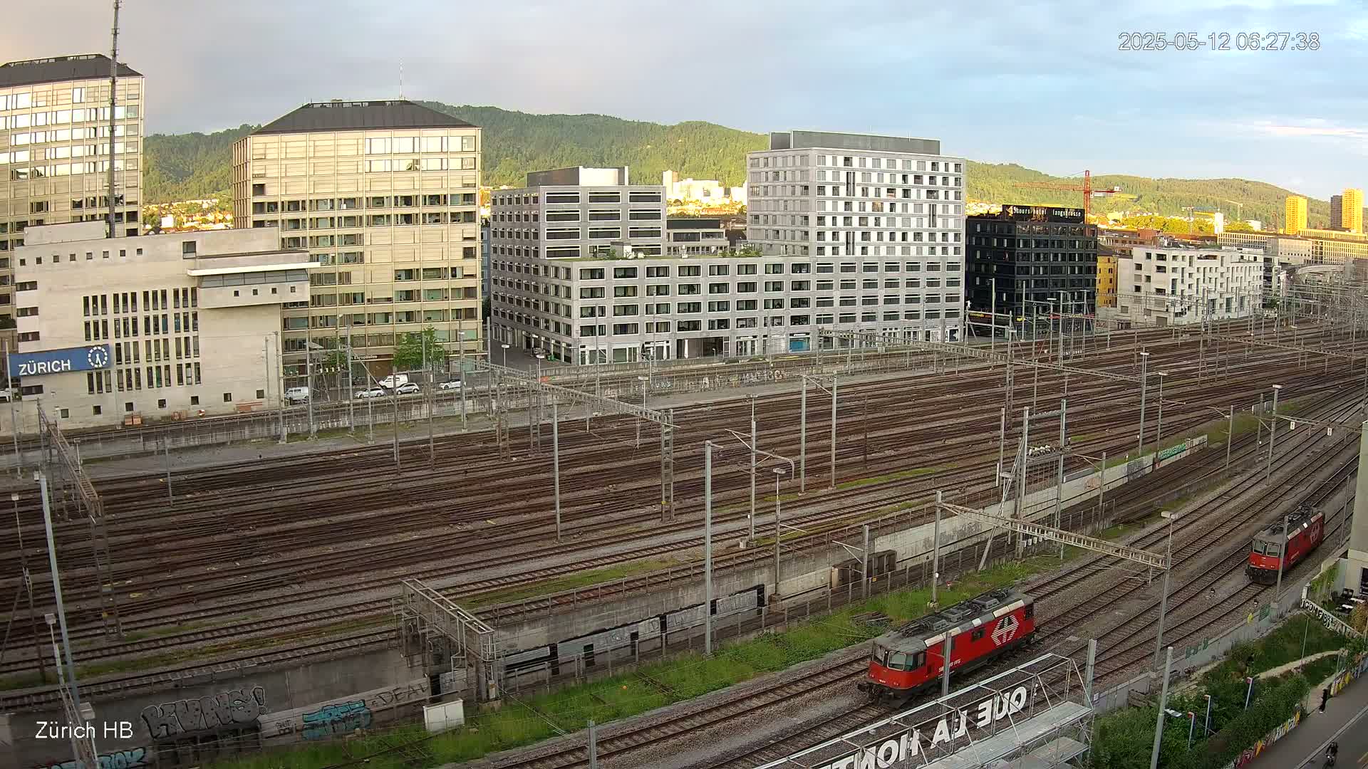 Two red locomotives are visible on a vast network of train tracks, alongside several modern buildings under a partly cloudy sky.