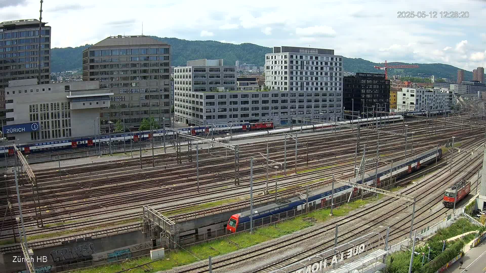 A high-angle view of a train station with multiple train tracks, several modern buildings, and a partly cloudy sky.
