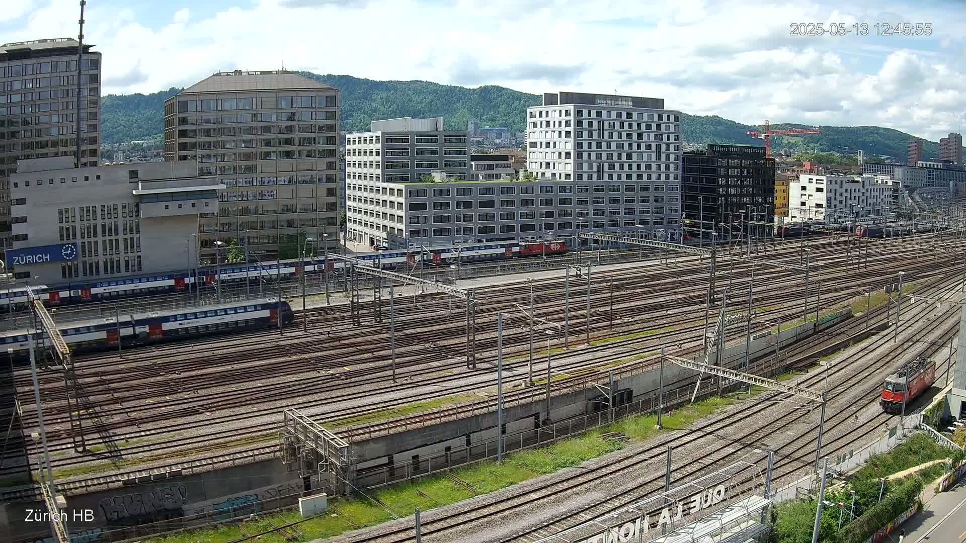 A sprawling railway yard filled with numerous train tracks, several trains, and modern buildings under a partly cloudy sky.