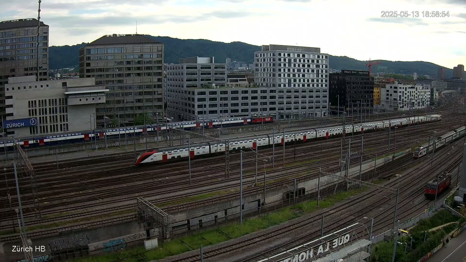 A long white and red train travels across a large rail yard next to several modern buildings under a partly cloudy sky.