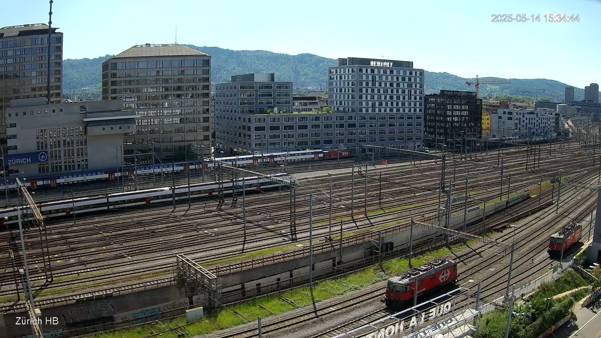 A sunny day view of a large railway yard with numerous train tracks, several passenger trains, two red locomotives, and a background of modern buildings and hills.