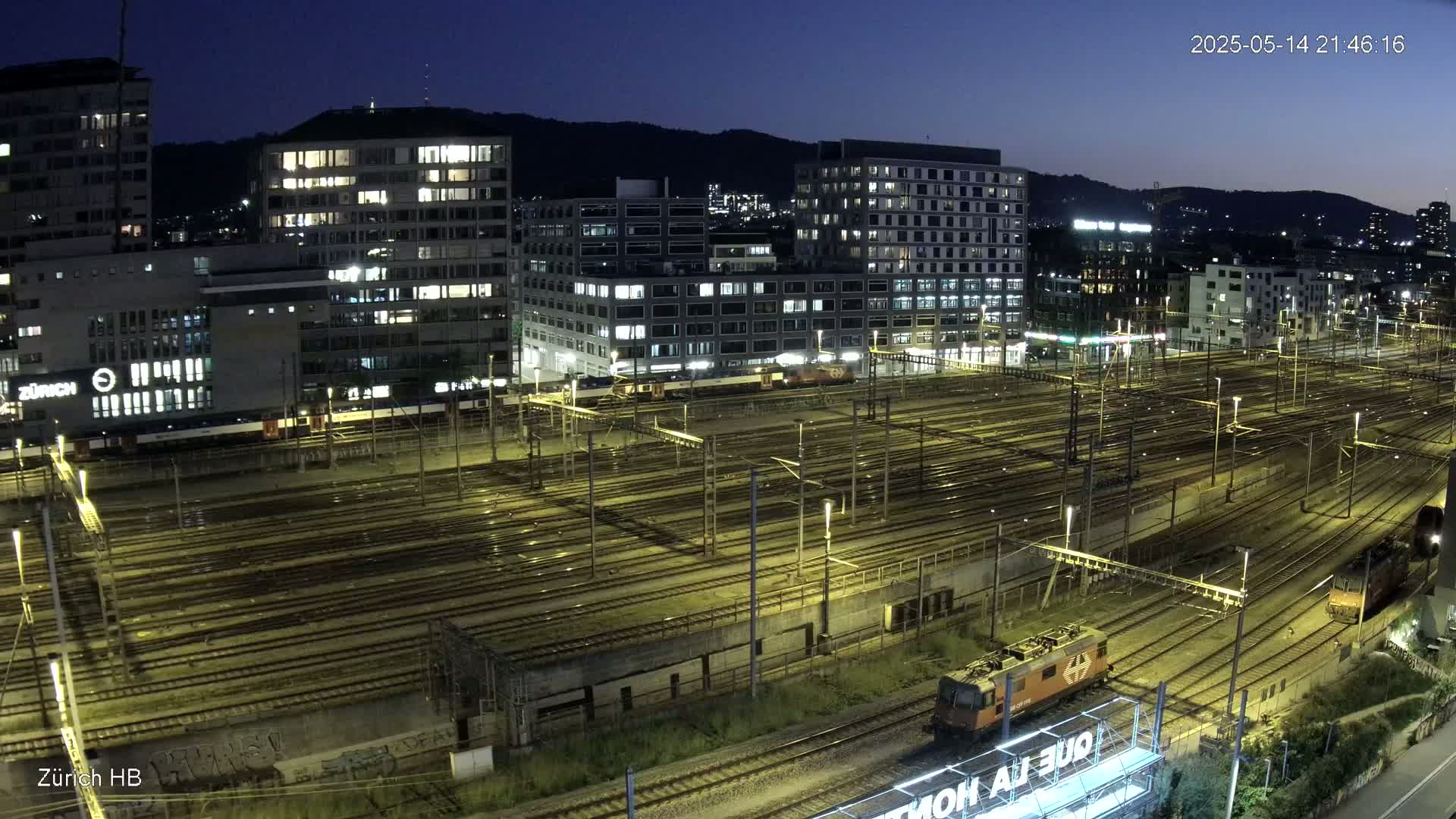 A nighttime view of a large, illuminated train yard with many tracks and several buildings in the background under a clear, dark sky.