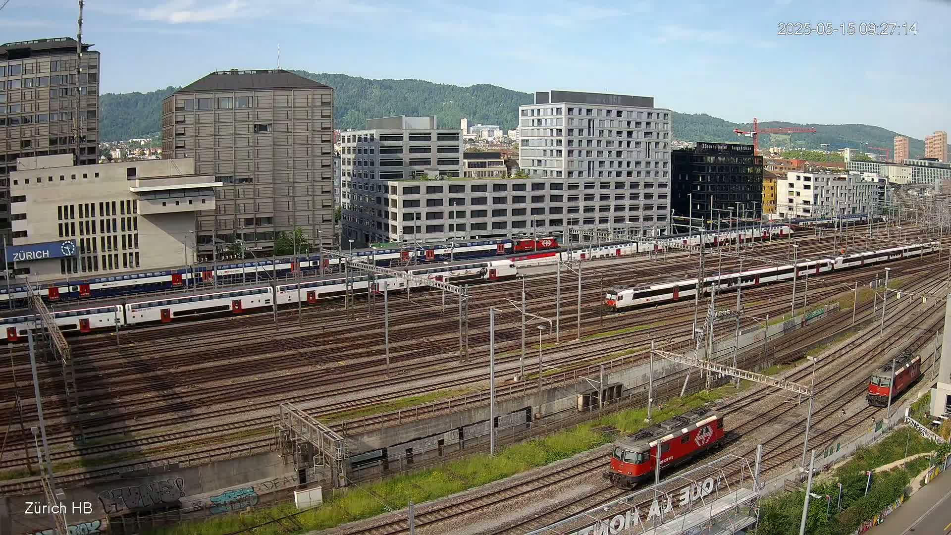 A sunny day overlooking a sprawling railway yard filled with numerous trains and surrounded by modern buildings and hills in the distance.