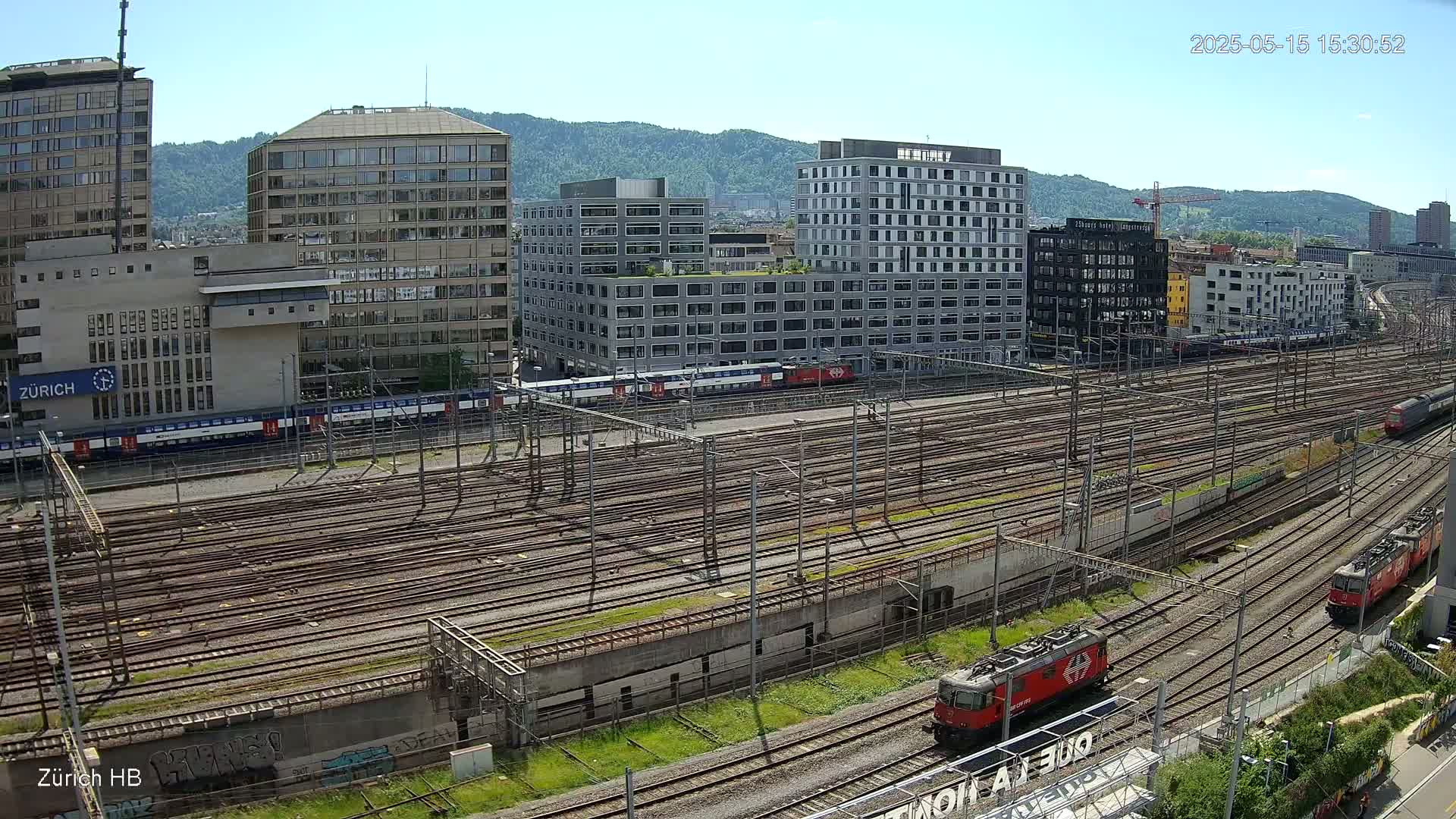 A sprawling railway yard with numerous tracks, several trains, and a backdrop of modern and older buildings under a clear, sunny sky.