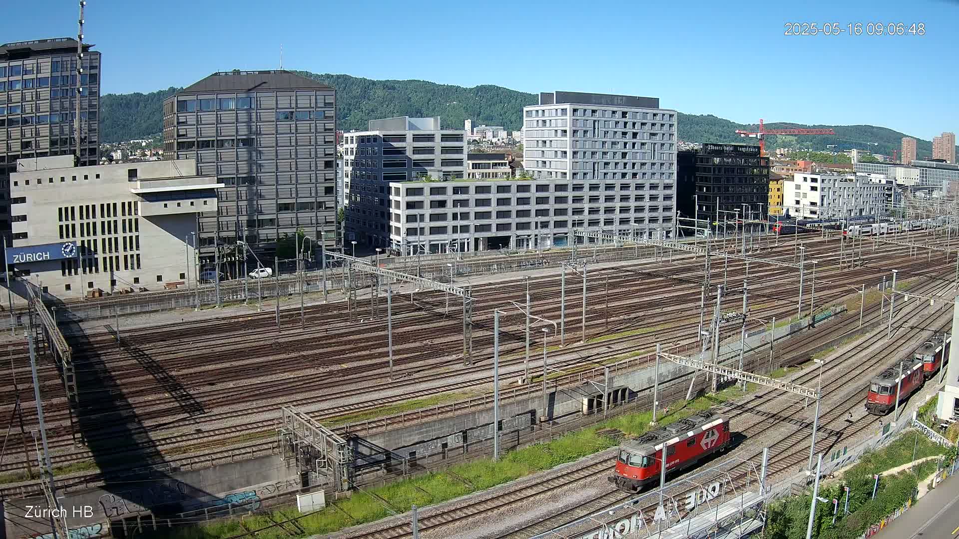 A sprawling railway yard with numerous tracks and two locomotives, situated amongst modern buildings on a clear, sunny day.